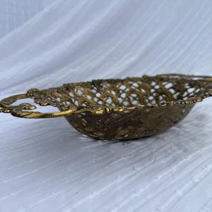 May include: A vintage, ornate brass bowl with a pierced, openwork design. The bowl has a shallow, oval shape with decorative handles on each side. The brass has a warm, aged patina, suggesting it is an antique or vintage piece. The bowl is likely used for decorative purposes.