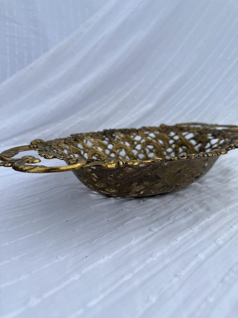 May include: A vintage, ornate brass bowl with a pierced, openwork design. The bowl has a shallow, oval shape with decorative handles on each side. The brass has a warm, aged patina, suggesting it is an antique or vintage piece. The bowl is likely used for decorative purposes.