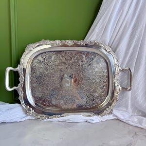 Puede incluir: Una bandeja de servicio rectangular de tono plateado con patrones florales y de volutas en relieve en su superficie. La bandeja incluye dos asas laterales y un borde decorativo festoneado con motivos en forma de concha en las esquinas.