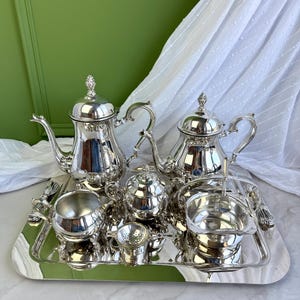 May include: A polished silver tea set consisting of a large teapot, a smaller teapot, a lidded sugar bowl, a creamer, a small tea strainer, and a bowl with a handle, all arranged on a rectangular silver serving tray with ornate feet.
