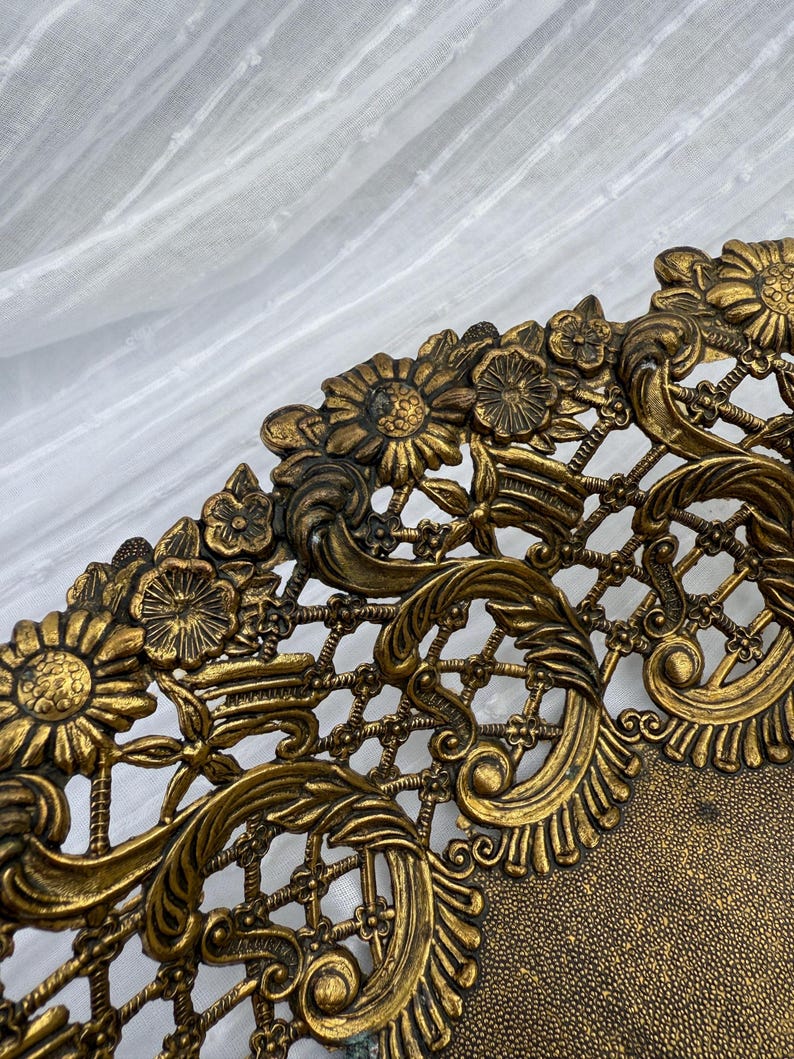 May include: Close-up of a decorative, antique-style tray with intricate floral and scrollwork details. The tray is made of a gold-toned metal and features a lattice design. The edges are adorned with detailed flowers and leaves.