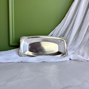 May include: A rectangular silver-toned metal tray featuring a polished, reflective basin and ornate, embossed floral detailing along the four corners and edges. The tray has a beaded border design and a smooth, slightly concave centre surface.