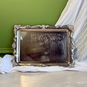 May include: A rectangular silver-coloured serving tray featuring an ornate, raised border with intricate scrollwork and shell-like motifs. The central surface of the tray is smooth and reflective, showcasing a subtle, etched floral pattern.