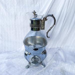 May include: A vintage silver-plated coffee or tea server with a clear glass carafe. The server has a decorative handle and base, and a small burner area. The glass carafe is approximately 20 cm tall.