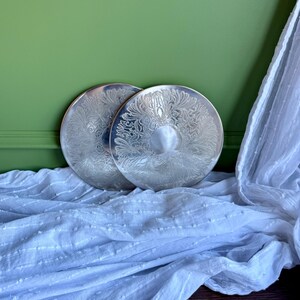 Può includere: Due piatti circolari in metallo color argento caratterizzati da un intricato motivo floreale e a foglie in rilievo che circonda un centro liscio e semplice. I piatti hanno una finitura lucida e riflettente e sono sovrapposti.