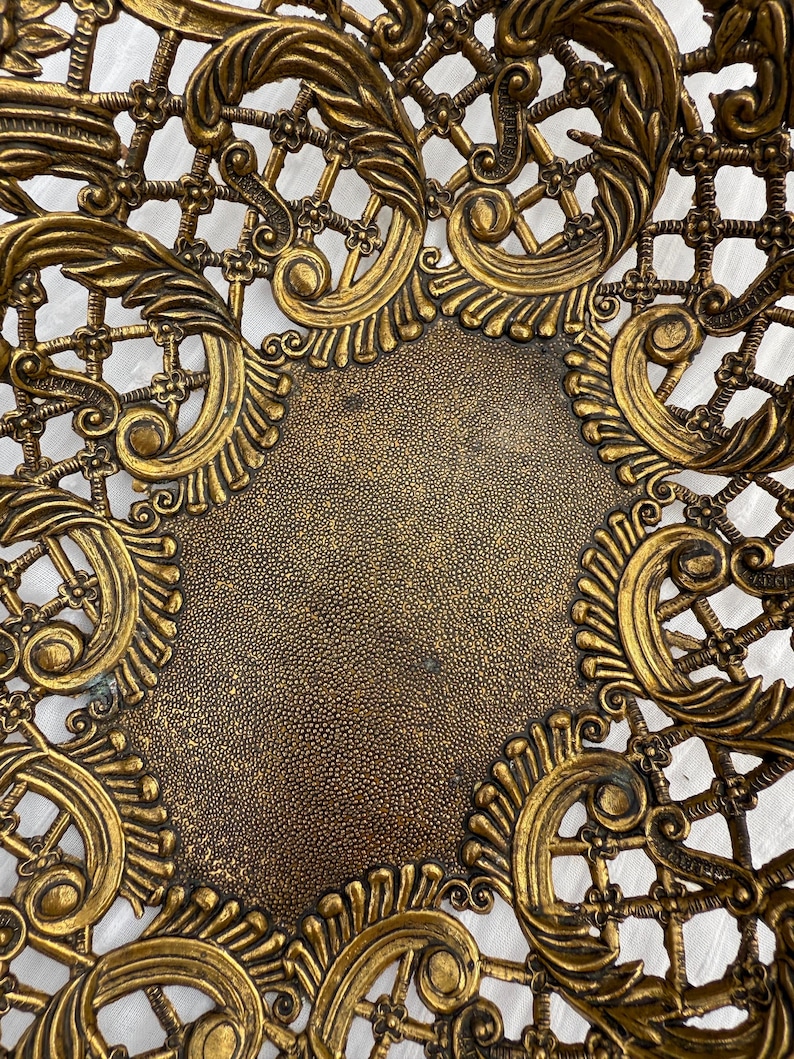 May include: Ornate, antique-style metal basket with a gold finish. The basket features a detailed openwork design with scrolling leaf and floral motifs. The center has a textured, solid surface. The overall aesthetic is vintage and decorative.