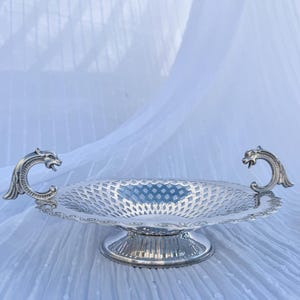 May include: Silver fruit bowl with a pierced design. The bowl has a round base and two dragon-shaped handles. The bowl is set against a white, textured background.