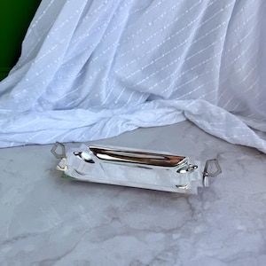 May include: A rectangular silver-toned metal tray featuring a polished, reflective surface and a recessed centre. The tray includes two geometric, diamond-shaped handles on opposite ends, providing a sleek and functional design for serving.