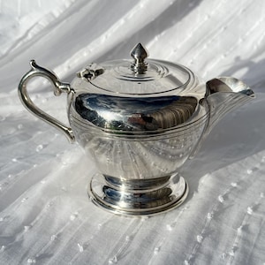 May include: A shiny silver teapot with a curved handle, spout, and a lid with a pointed finial. The teapot sits on a round base and reflects the surrounding environment. The teapot is likely made of metal.