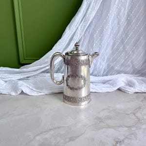 May include: A silver-toned metal coffee pot featuring a cylindrical body with an ornate embossed floral band around the top and bottom, a central oval decorative frame, a curved handle, a hinged lid with a small finial, and a short spout.