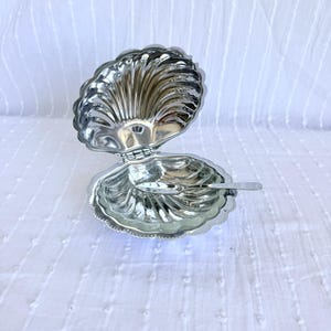 May include: A silver-plated shell-shaped serving dish with a hinged lid and a small spoon. The dish is open, revealing its interior, and sits on a white, textured surface. The shell design is detailed and ornate.