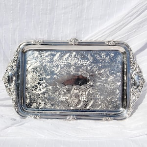 May include: A rectangular silver serving tray with ornate floral and scrollwork detailing. The tray has decorative handles on each end and a raised rim. The interior features a checkered pattern and a reflective surface.