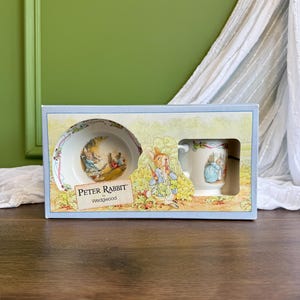 Puede incluir: Un juego de cuenco y taza de cerámica Peter Rabbit de Wedgwood en una caja de cartón rectangular. El empaque presenta ilustraciones de Peter Rabbit en un jardín, con el texto Peter Rabbit by Wedgwood impreso en el frente de la caja.