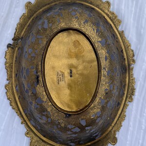 May include: An antique brass oval-shaped decorative bowl with ornate filigree detailing. The bowl has two handles and a decorative rim. The bottom of the bowl has an oval-shaped gold center. The words "Made in Italy" are visible.