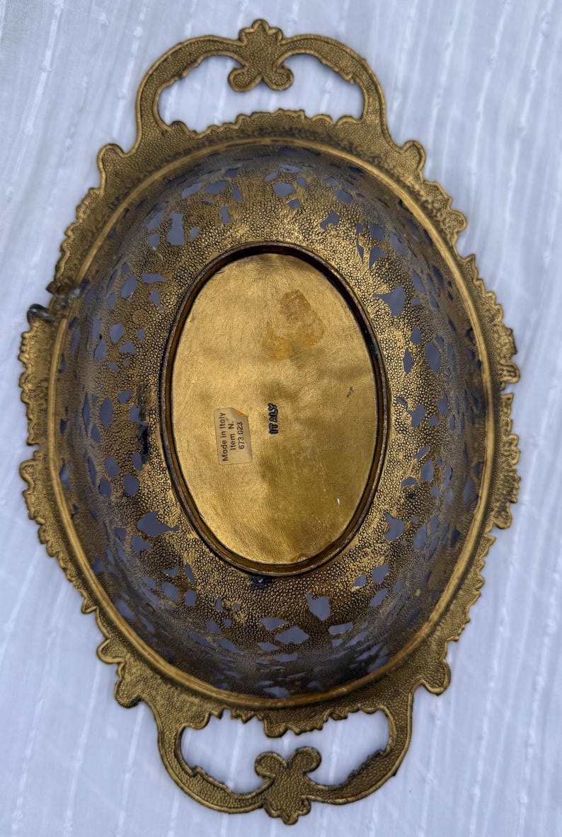 May include: An antique brass oval-shaped decorative bowl with ornate filigree detailing. The bowl has two handles and a decorative rim. The bottom of the bowl has an oval-shaped gold center. The words "Made in Italy" are visible.