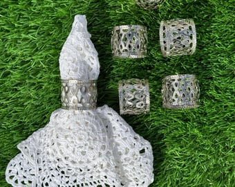 May include: Silver metal napkin rings with an openwork design, alongside a white crocheted napkin on a green surface. The napkin rings feature a decorative pattern. The items are arranged in a group.