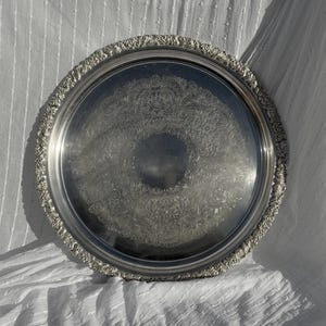 May include: A circular, silver-coloured serving tray with an elaborate floral design in the centre and around the edge. The tray is made of metal and has a reflective surface. The tray is on a white, textured surface.