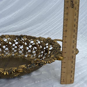 May include: Ornate gold-colored metal basket with a pierced floral design. The basket has handles and measures approximately 8 inches long. The basket is on a white background.