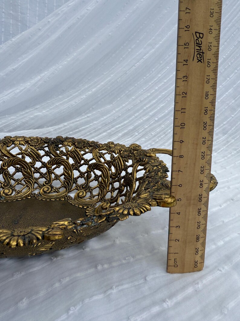 May include: Ornate gold-colored metal basket with a pierced floral design. The basket has handles and measures approximately 8 inches long. The basket is on a white background.