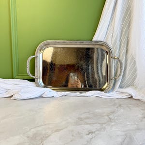 Puede incluir: Una bandeja de servicio rectangular de tono plateado con esquinas redondeadas, que presenta un patrón floral en relieve en la base, un borde con cuentas y dos asas curvas en los lados. La superficie pulida refleja el entorno.