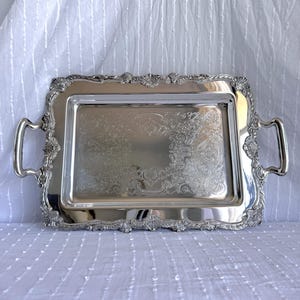 Puede incluir: Una bandeja rectangular plateada con detalles ornamentados. La bandeja presenta un patrón floral decorativo en el centro y a lo largo de los bordes. Tiene dos asas curvas a cada lado.