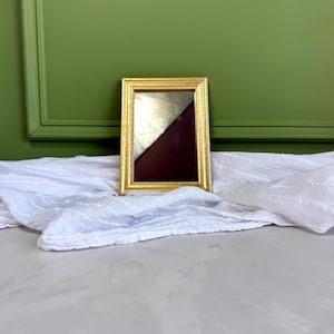 May include: A rectangular mirror featuring a thin, ornate gold-coloured frame with a beaded inner edge. The mirror surface reflects a dark, angled shadow, and the frame has a textured, metallic finish that catches the light along its edges.