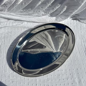 May include: Oval, silver-coloured serving tray with a polished finish. The tray has a raised, rounded edge and reflects the surrounding environment. The tray is set against a white, textured background.