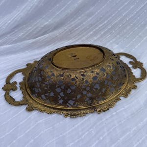 May include: An antique gold-toned metal decorative bowl with intricate cut-out patterns. The oval-shaped bowl features ornate handles on each side and a detailed, scalloped edge. The interior has a dark, possibly painted, surface.