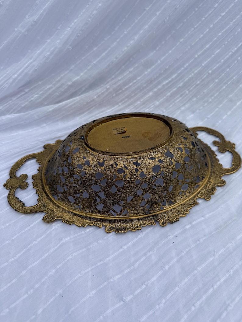 May include: An antique gold-toned metal decorative bowl with intricate cut-out patterns. The oval-shaped bowl features ornate handles on each side and a detailed, scalloped edge. The interior has a dark, possibly painted, surface.