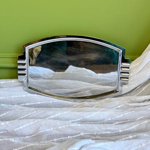 May include: A rectangular silver-toned metal tray with a polished, reflective surface. The tray features curved top and bottom edges and decorative stepped, ribbed detailing on the left and right sides, creating a structured, symmetrical appearance.