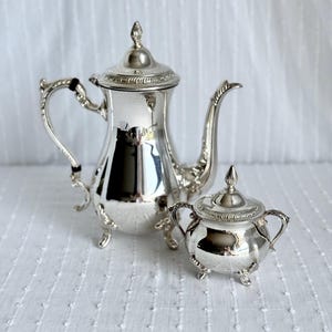 Puede incluir: Un juego de té plateado con una tetera y un azucarero. La tetera tiene un pico curvo y un asa con un acento negro. El azucarero tiene dos asas y una tapa. Ambas piezas tienen detalles ornamentados y están sobre pequeños pies.