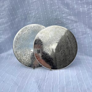 May include: Two silver-colored, round decorative plates with intricate floral patterns. The plates are displayed on a white, textured surface. The plates have a reflective surface, and the floral design is etched into the metal.