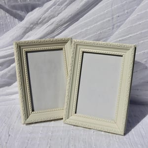 May include: Two cream-coloured rectangular picture frames with decorative scrollwork. The frames are empty, with a white interior, and are set against a white, textured background. The frames are slightly angled.