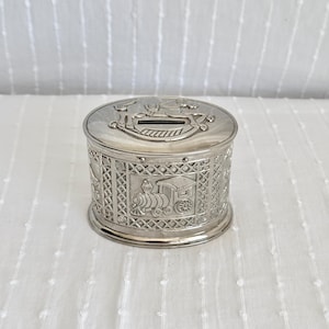 May include: A silver-coloured, cylindrical money box with detailed embossed designs. The top has a slot for inserting money and a rocking horse motif. The sides display intricate patterns and a train carriage scene, creating a vintage aesthetic.