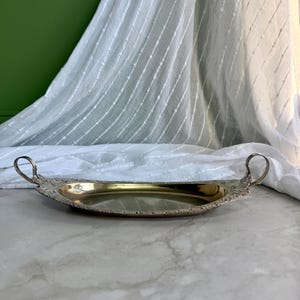 May include: An oval-shaped brass serving tray featuring a polished gold-toned interior basin and an ornate, textured silver-toned rim. The tray includes two curved handles on each end, decorated with intricate floral and scrollwork detailing.