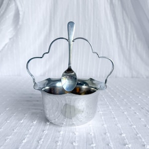 May include: A silver sugar bowl with a matching spoon. The bowl has a scalloped rim and decorative handles. The spoon rests on the handles. The bowl and spoon are made of a shiny metal.