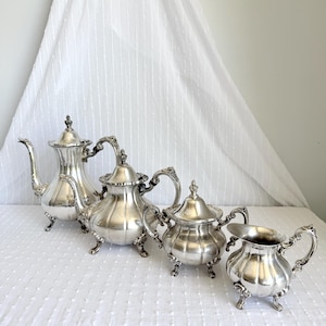 May include: A silver tea set comprising a teapot, a sugar bowl with a lid, a creamer jug, and a second teapot. The set features decorative handles and rests on small feet. The items are arranged on a white surface.