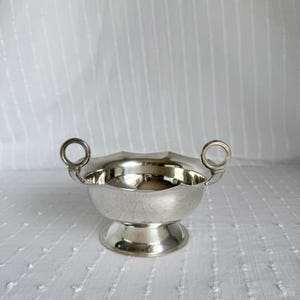 Puede incluir: Un pequeño cuenco plateado con borde festoneado y dos asas de anillo. El cuenco se asienta sobre una base de pedestal. El interior del cuenco refleja el entorno. El cuenco es probablemente de metal.