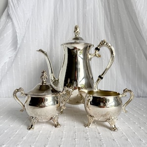 Puede incluir: Un juego de té plateado con tetera, azucarero y lechera. El juego tiene asas y patas ornamentadas. La tetera tiene una tapa con un adorno decorativo. El juego se exhibe sobre una superficie blanca.