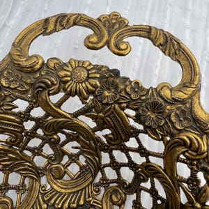May include: Ornate gold-colored metal tray with a floral and scroll design. The tray features a handle and intricate openwork detailing, including flowers and a lattice pattern. The overall aesthetic is vintage and decorative.