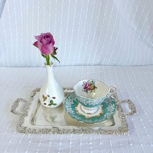 Puede incluir: Una bandeja plateada con bordes y asas ornamentadas sostiene un jarrón blanco con una rosa rosa, y una taza y platillo. La taza y el platillo tienen un diseño turquesa y floral. El conjunto está sobre una superficie blanca texturizada.