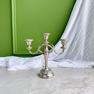 May include: A silver-toned metal candelabra featuring three candle holders with a central stem and two curved arms. The base and the top of each candle cup are decorated with a raised floral pattern, and the structure has a polished finish.
