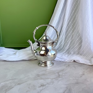May include: A polished silver-toned teapot featuring a rounded body, a pedestal base, and a curved spout. The teapot includes a hinged lid with a small finial and an arched handle with ornate detailing at the connection points to the main body.