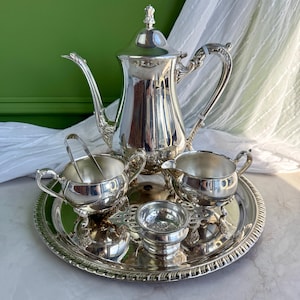 May include: A silver-plated tea service set consisting of a tall teapot with a lid, a creamer, a sugar bowl with tongs, and a small tea strainer, all arranged on a round, ornate silver tray with a decorative scalloped edge and polished finish.