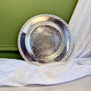 May include: A round silver-toned metal serving tray featuring an intricate embossed floral pattern along the wide rim and a central area with a polished finish. The tray has a decorative scalloped edge and a reflective surface showing light reflections.