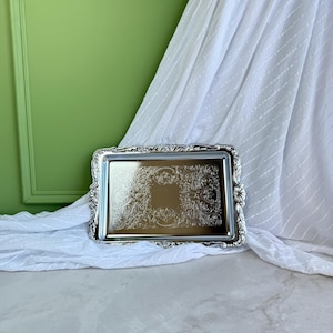 Puede incluir: Una bandeja de servicio rectangular de tono plateado con un borde ornamentado y repujado de motivos florales y volutas. La superficie central de la bandeja es lisa y reflectante, enmarcada por intrincados detalles metálicos.