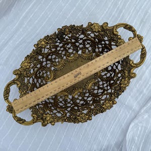 May include: An oval, antique-style brass tray with floral and scrollwork detailing. The tray has two handles and measures approximately 12 inches long. The openwork design is accented with small flowers around the edges.