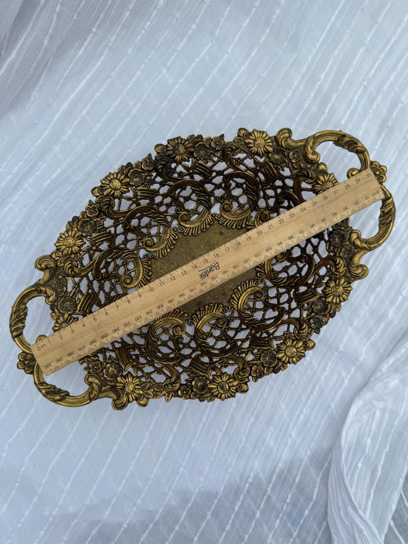 May include: An oval, antique-style brass tray with floral and scrollwork detailing. The tray has two handles and measures approximately 12 inches long. The openwork design is accented with small flowers around the edges.