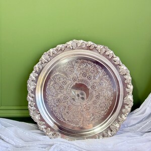 May include: A round silver-toned metal serving tray featuring an intricate embossed floral and scrollwork pattern in the centre. The raised outer rim is decorated with a detailed relief design of grape clusters and vine leaves along the edge.
