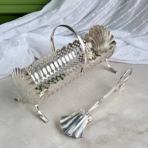 May include: A silver-toned metal basket with a lattice-patterned body, a curved handle, and shell-shaped ends, supported by four small legs. Accompanying the basket is a matching silver-toned metal serving utensil with a fan-shaped end and a ring handle.